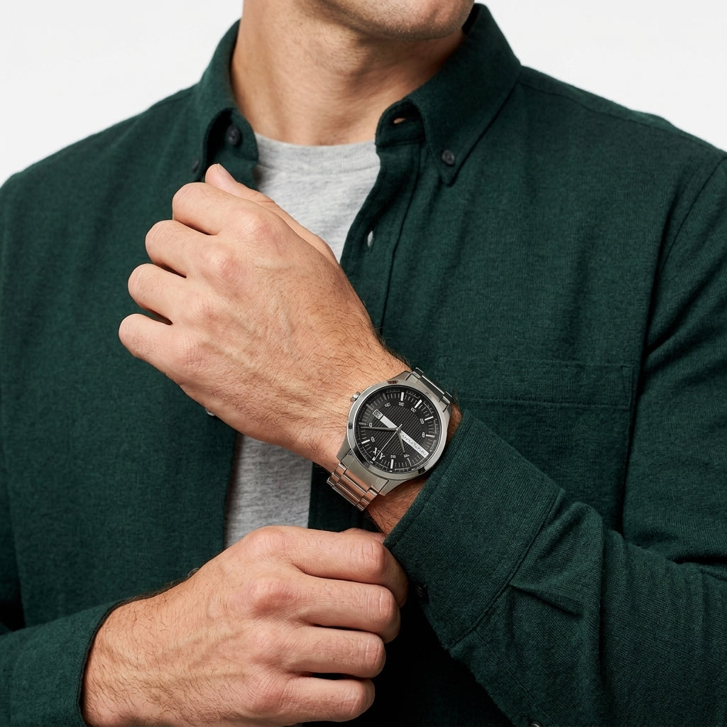 A close-up of a man wearing a silver stainless steel watch with a black dial and a matching metal link bracelet, styled with a dark green button-down shirt over a light gray t-shirt.